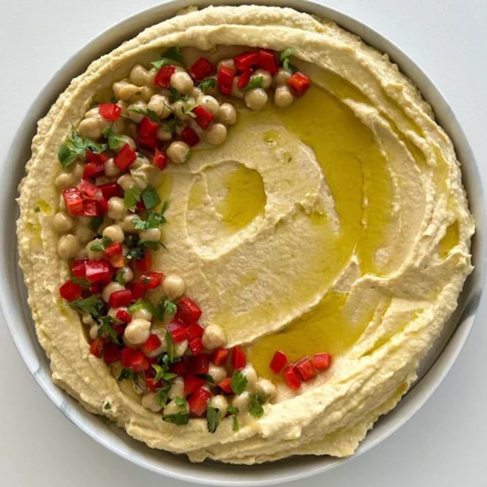 Creamy hummus topped with chickpeas, diced red peppers, fresh herbs, and olive oil in a white bowl.