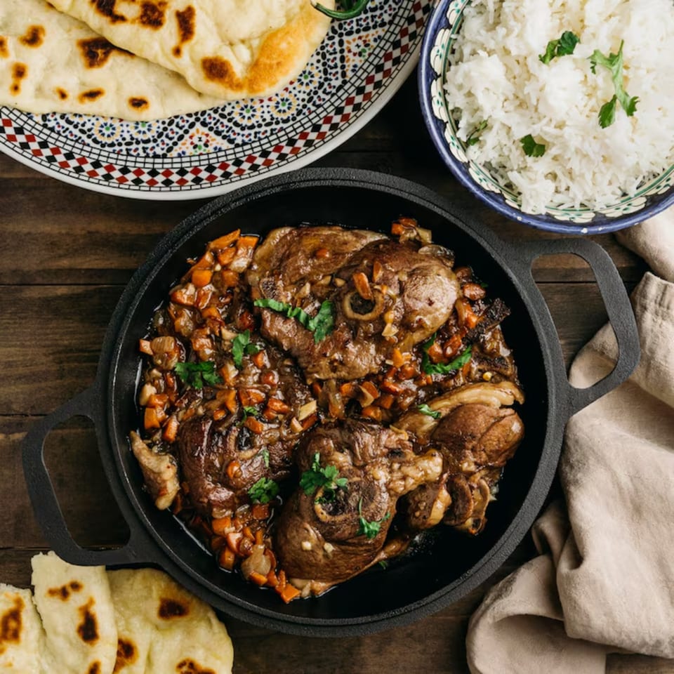 A cast iron skillet of braised lamb shanks with carrots, served with rice and naan bread.