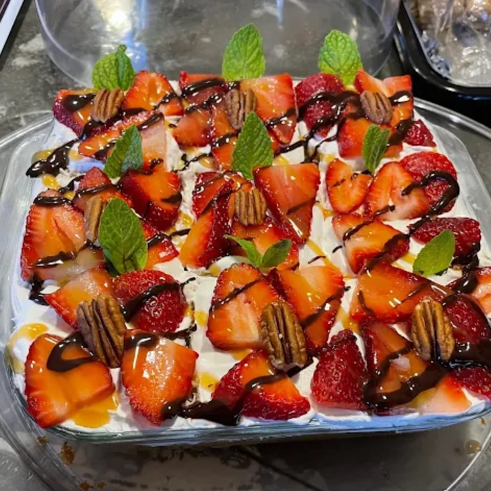 A square dessert with whipped cream, strawberries, pecans, chocolate, and mint leaves.