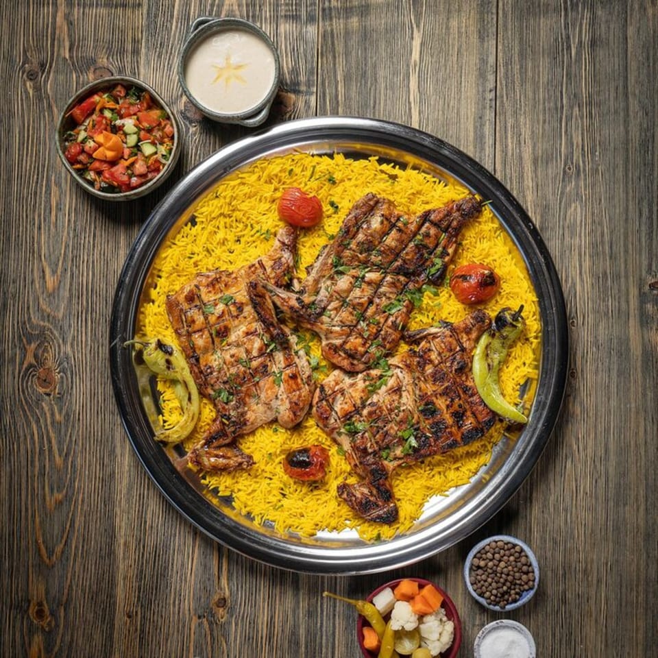 Grilled chicken pieces on yellow rice with roasted vegetables, salad, and sauces on a wooden table.