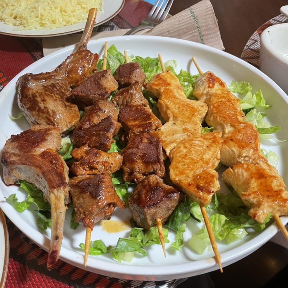 A white plate of Mediterranean lamb chops, beef skewers, and chicken skewers on a bed of lettuce.